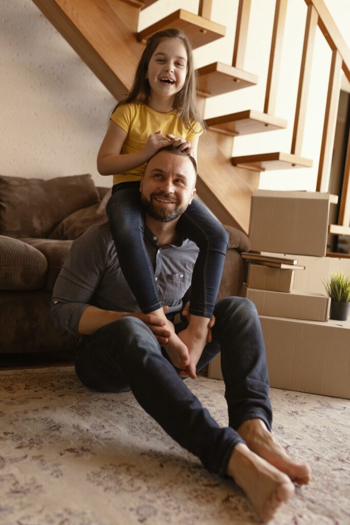 Un père monoparental, vêtu d'un t-shirt décontracté, sourit tandis qu'il porte sa fille sur ses épaules. Sa fille rit joyeusement, ses bras enlacés autour du cou de son père.